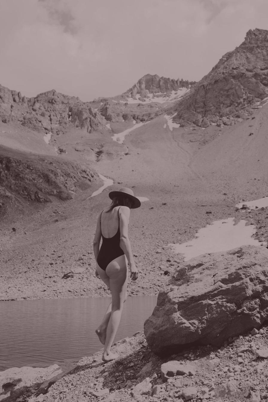A woman standing alone at the edge of an alpine lake, surrounded by open mountains