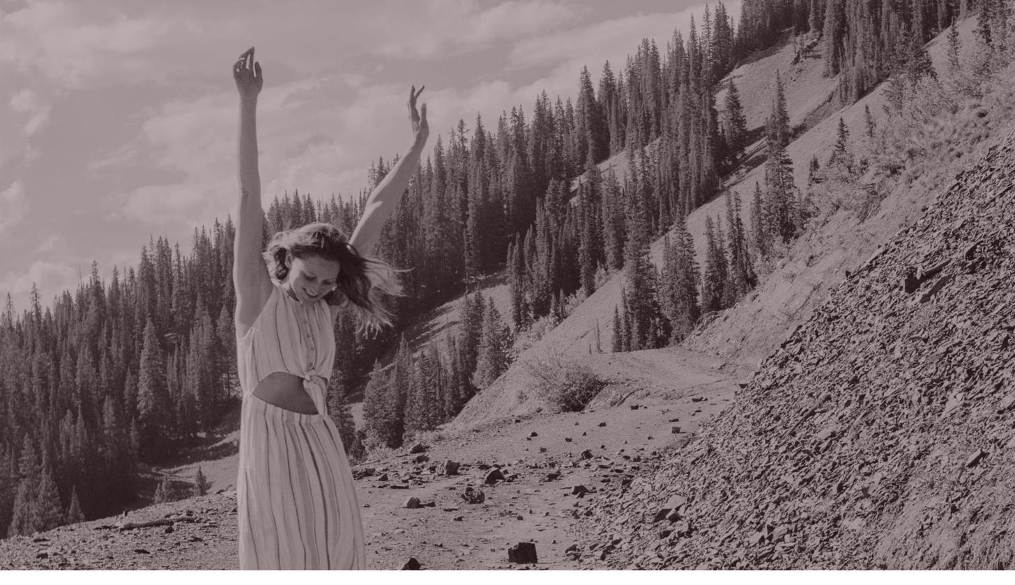Rita with arms raised on a mountain road, wind in her hair
