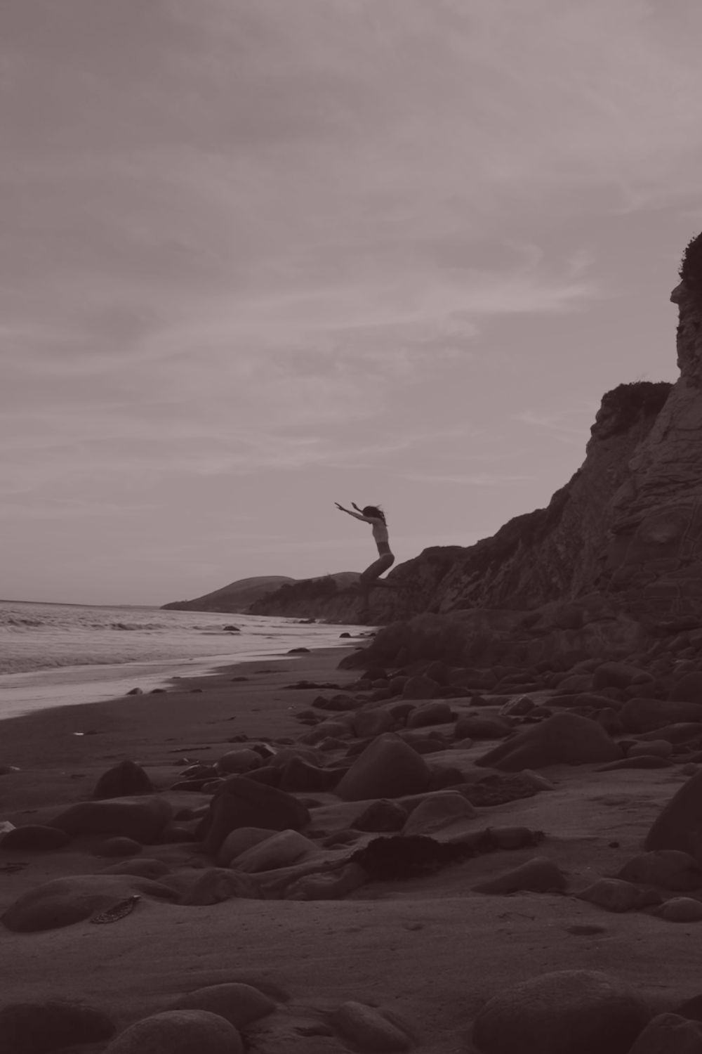 A woman leaping joyfully from a cliff toward the beach below, arms raised