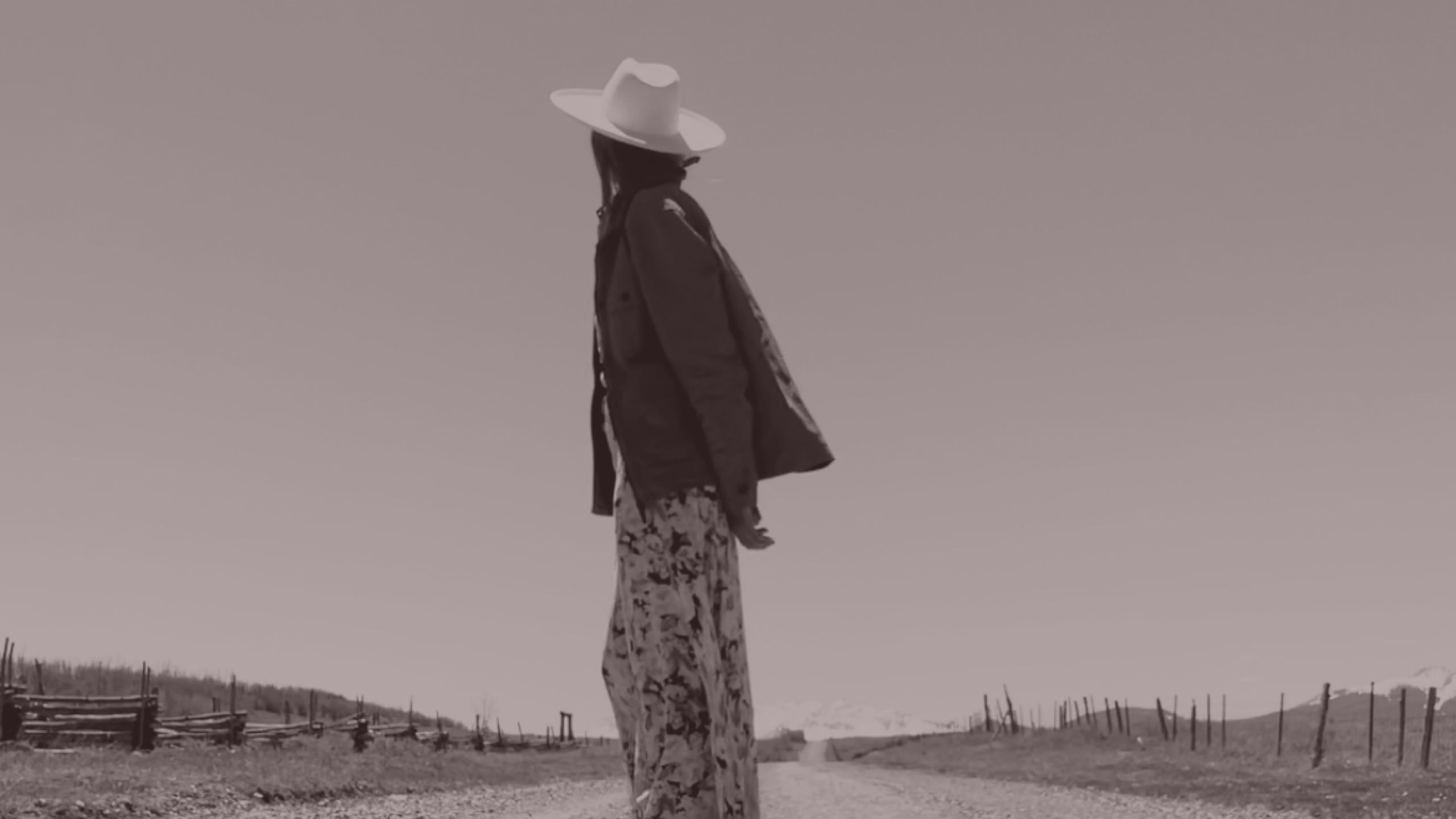 A woman in a white hat walking down a quiet dirt road toward distant mountains