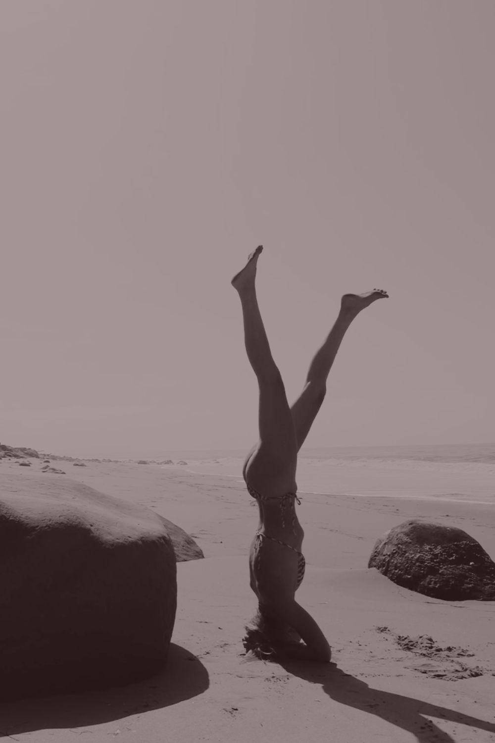 A woman in a headstand on the beach, legs open toward the sky