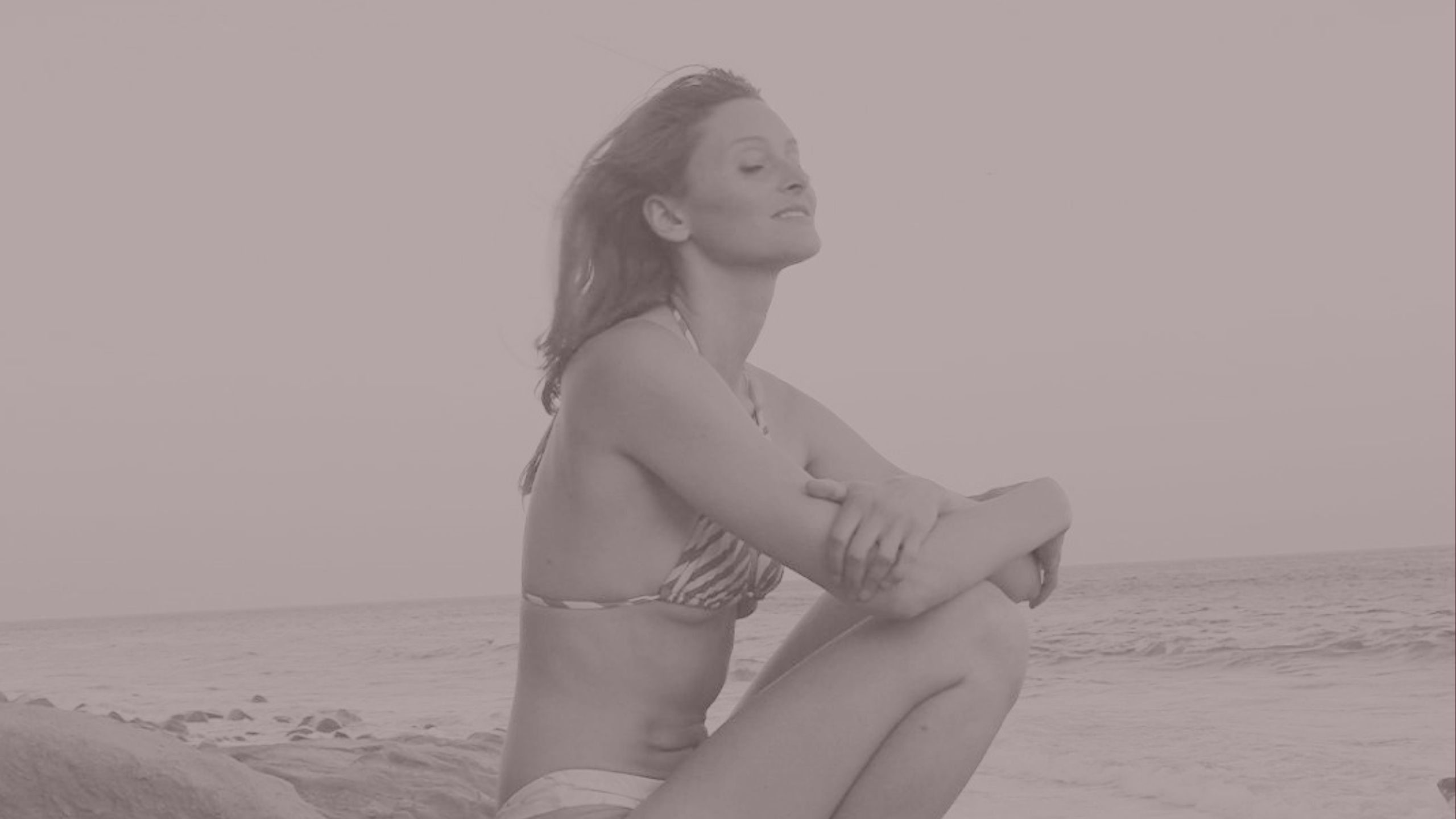 A woman sitting quietly on a rock at the ocean's edge, eyes closed, listening