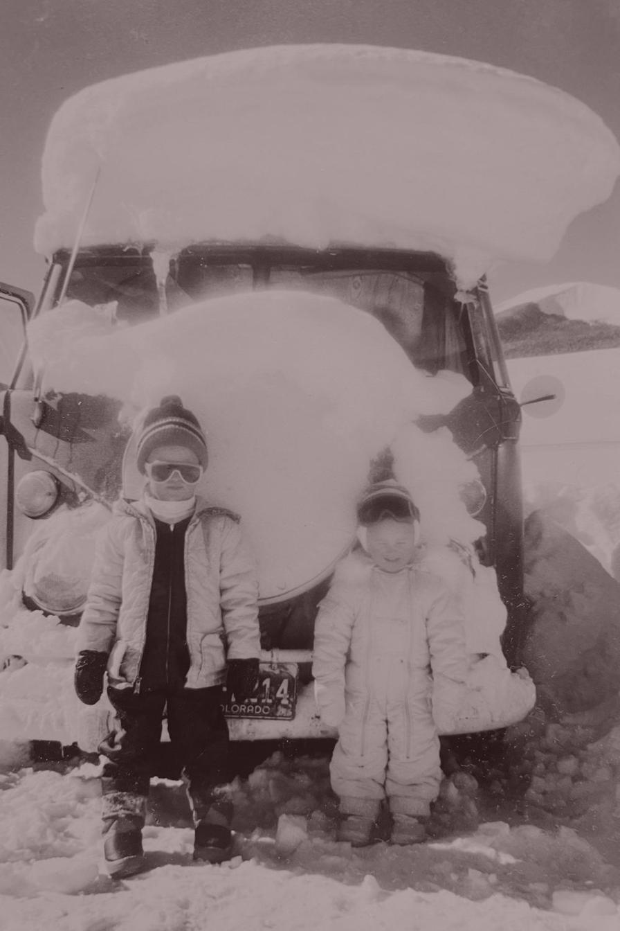 Two children bundled in snow gear in front of a truck buried in snow