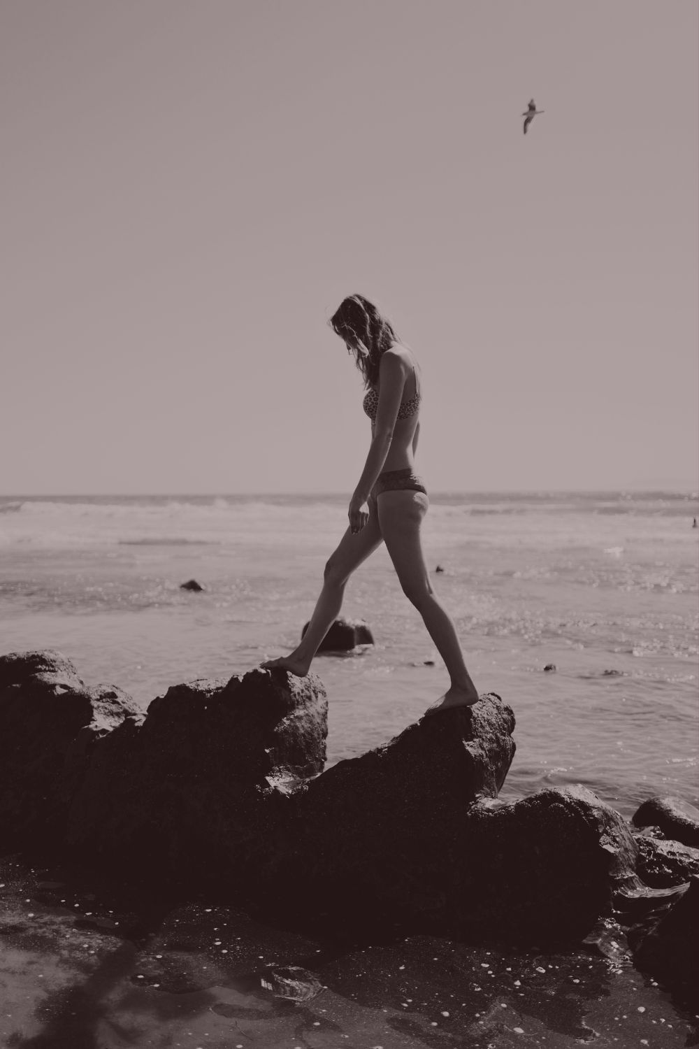A woman walking barefoot along ocean rocks, head bowed in quiet attention